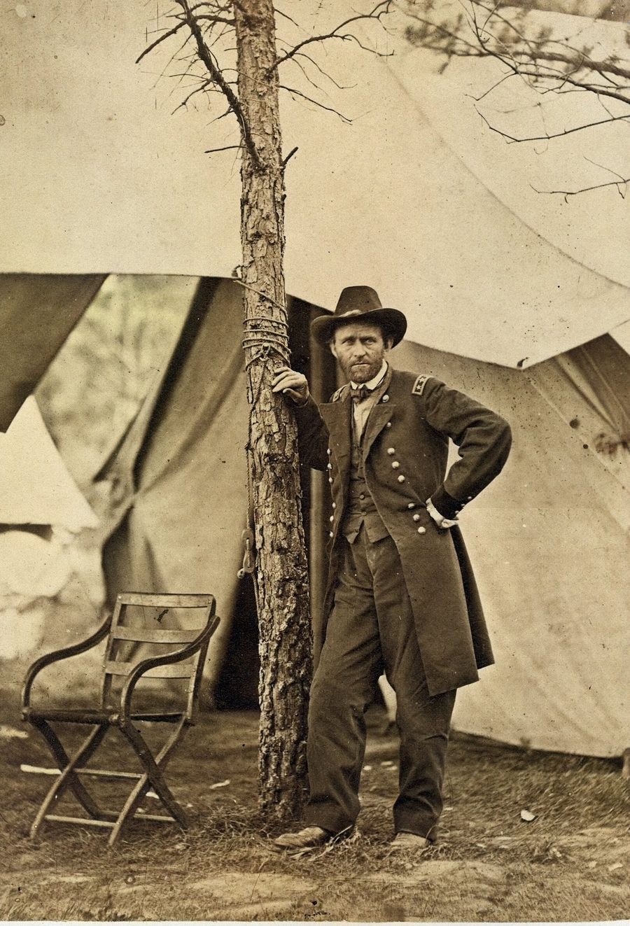 Colorized photograph of Grant in uniform and a slouch hat, leaning on a tree in front of a canvas army tent
