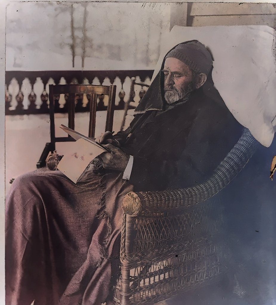 Colorized photograph of a thin, bearded Grant bundled in blankets and a knit cap, sitting in a wicker chair on a porch with papers in his lap
