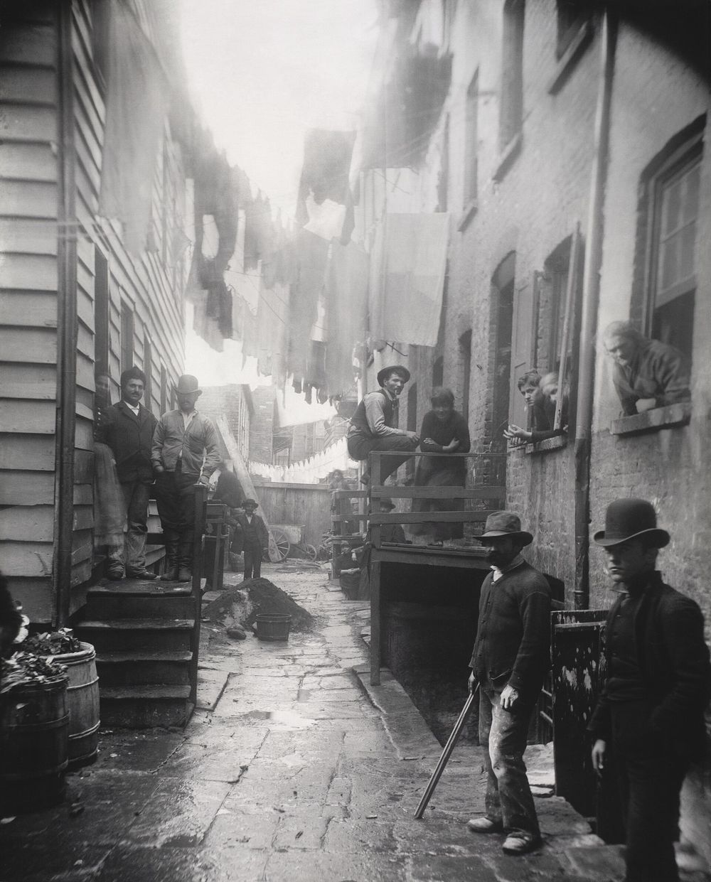 A narrow alley between tenement buildings with laundry hanging overhead and men in hats standing along the walls.