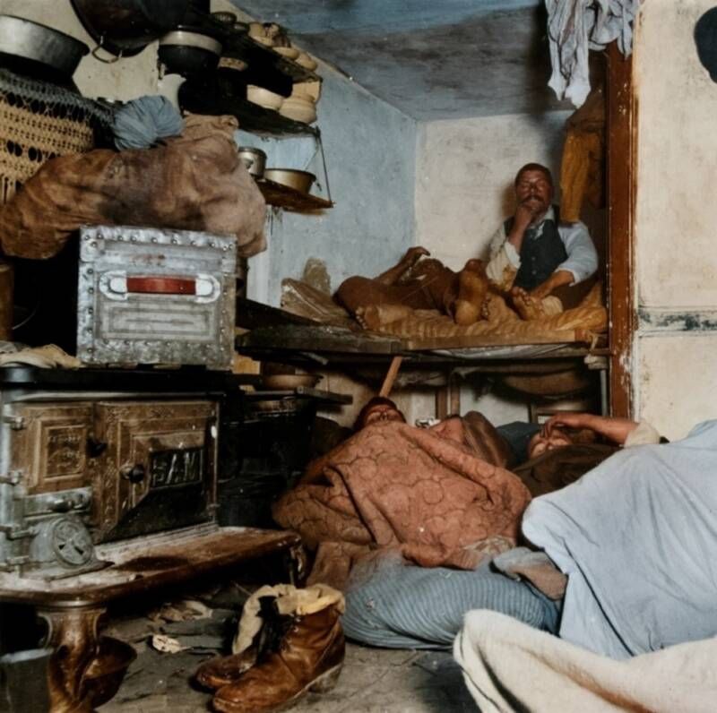 A cramped room with bunks and men sleeping among bundles and pots.