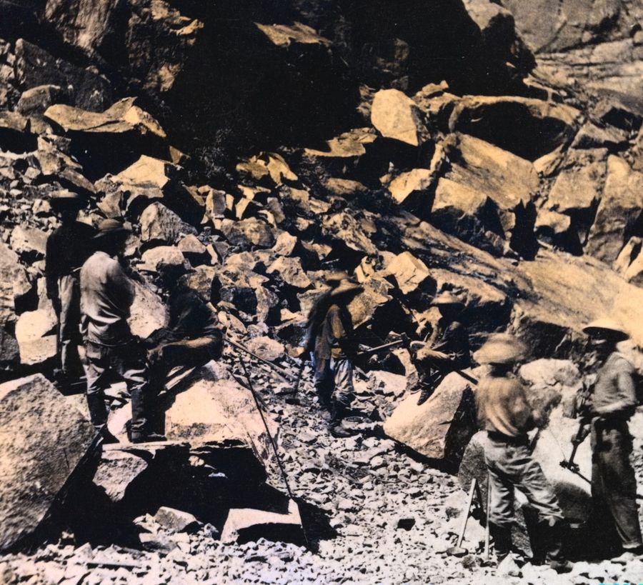 Chinese laborers in wide hats working among huge broken boulders on a mountainside.