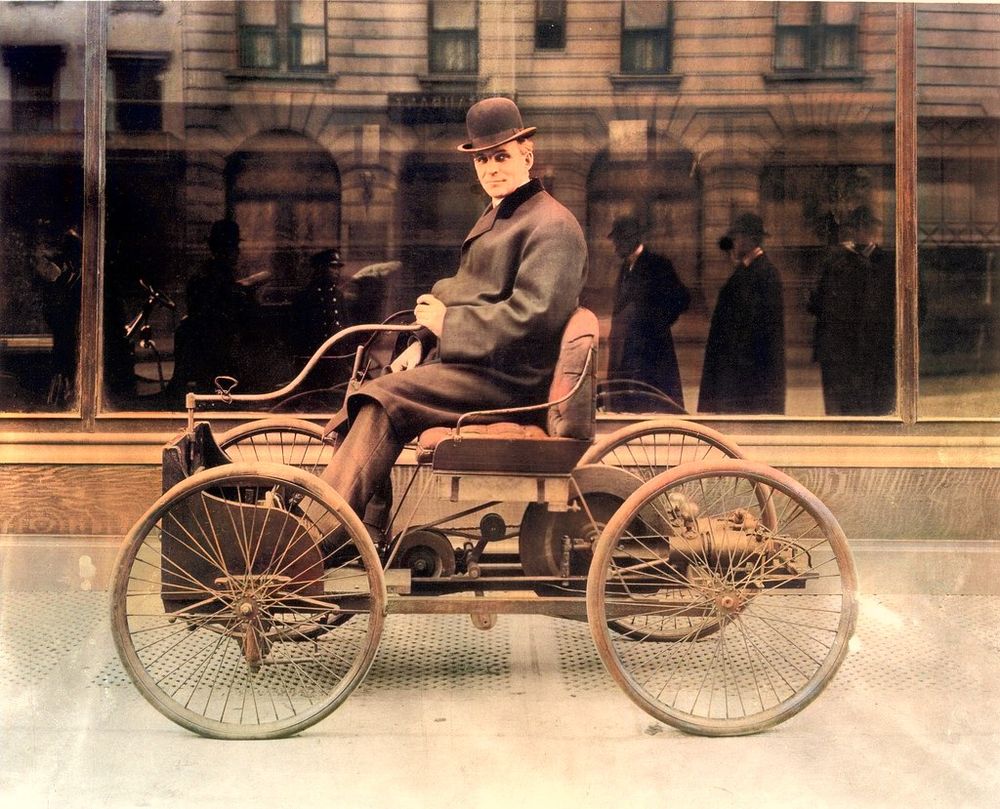 Henry Ford in a bowler hat seated in the Quadricycle, a small four wheeled car with thin bicycle wheels, in front of a building.