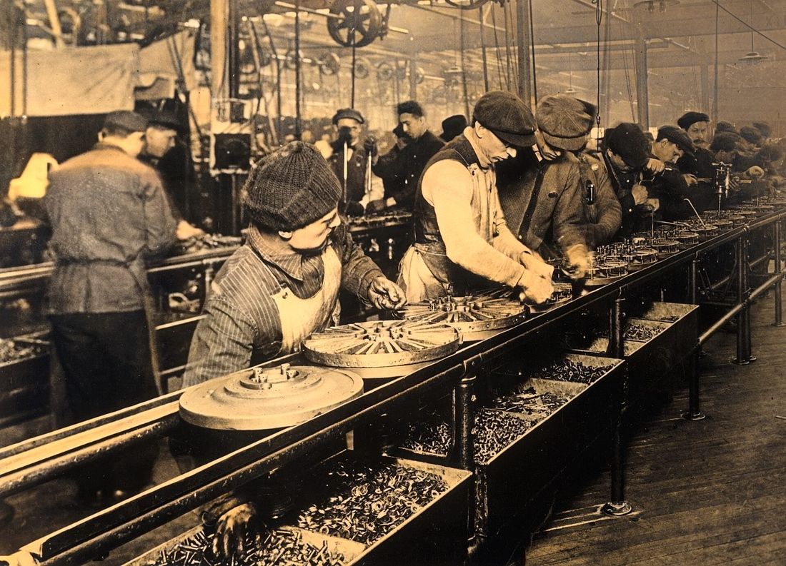 A row of workers in caps standing along a long bench assembling round flywheel parts.
